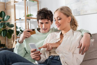 cheerful woman pointing at smartphone while doing online shopping near boyfriend holding credit card 