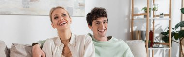 happy and curly young man hugging blonde girlfriend at home, banner 
