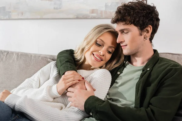 young and curly man hugging happy blonde girlfriend at home