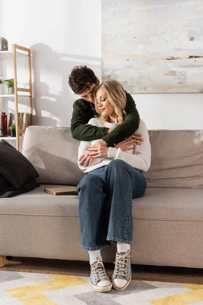 Curly Young Man Hugging Happy Blonde Woman Sitting Sofa Living — Stockfoto