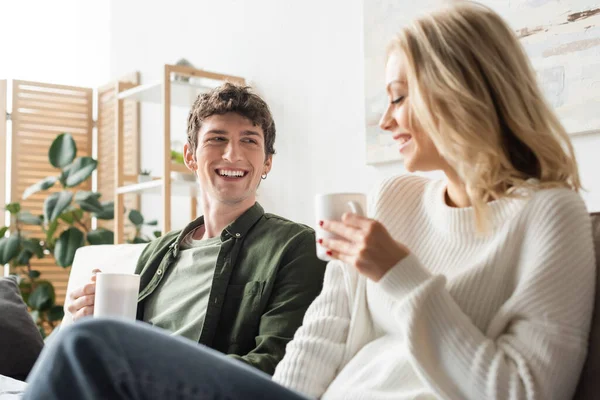 cheerful young man looking at blonde woman in sweater while holding cup with coffee in living room 