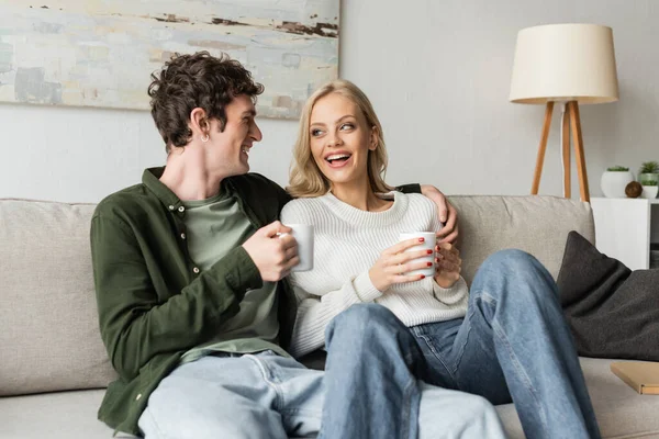 cheerful young man hugging blonde woman in sweater while holding cup with coffee in living room 