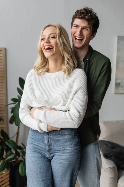 excited man with curly hair hugging girlfriend and laughing at home 