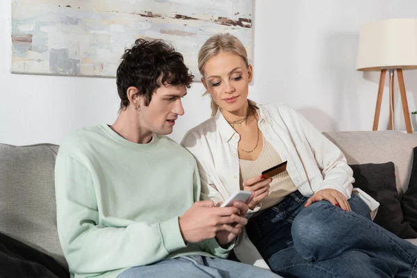blonde woman holding credit card near boyfriend with smartphone doing online shopping 