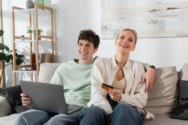 cheerful woman holding credit card near boyfriend with laptop doing online shopping 