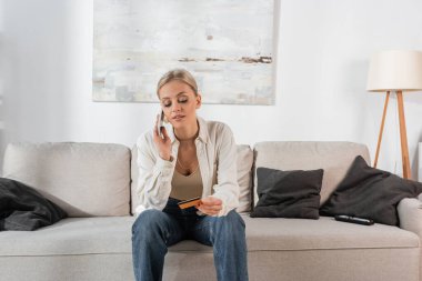 blonde woman holding credit card and talking on smartphone in living room 