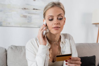 young blonde woman holding credit card while talking on smartphone in living room 