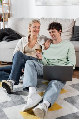 cheerful man holding credit card while using laptop near smiling girlfriend 