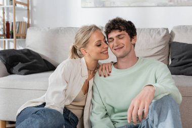 happy blonde woman with closed eyes smiling near boyfriend in living room 