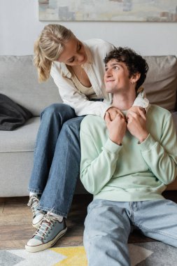 young and blonde woman embracing pleased man in living room 