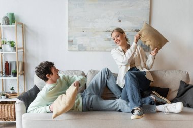 full length of happy young couple having pillow fight on couch in living room 