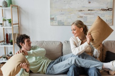 positive young couple smiling while having pillow fight in living room 