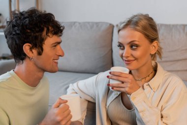 happy woman with blonde hair and curly man looking at each other while holding cups with coffee 