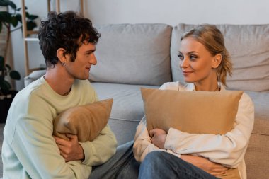 happy woman with blonde hair and curly man looking at each other while holding pillows 