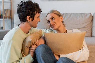 cheerful young woman with blonde hair and curly man looking at each other while holding pillows 