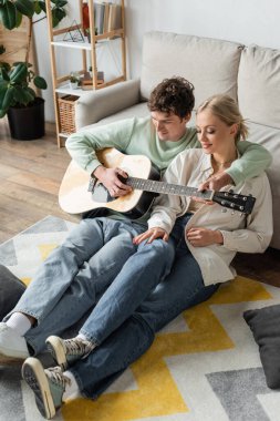 curly man playing acoustic guitar near blonde woman while sitting on carpet 