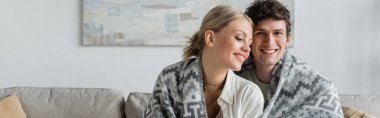 happy young man and woman covered in blanket smiling in living room, banner 