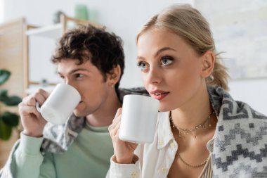 curly young man and blonde woman covered in blanket holding cups of tea 