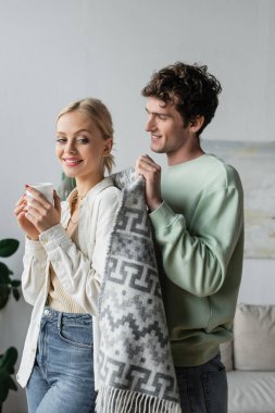 cheerful and caring man covering smiling blonde woman with blanket 