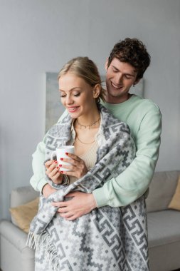 cheerful and caring man hugging blonde girlfriend covered in blanket and holding cup of tea 
