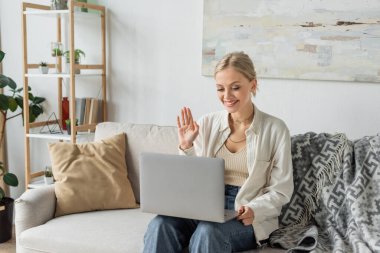 cheerful young woman with blonde hair waving hand during video call on laptop 