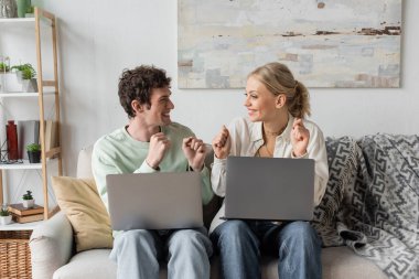 excited young bloggers rejoicing near laptops while sitting on couch  clipart