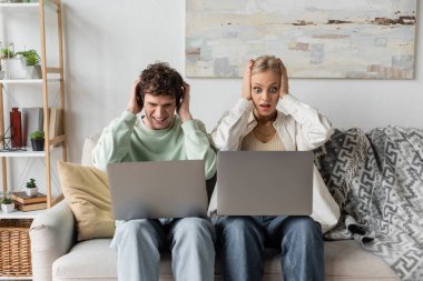 stressed young man and woman looking at laptops while sitting on couch 