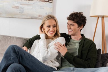 happy and curly man in shirt hugging pretty girlfriend in white sweater 