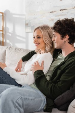 smiling and curly man in shirt hugging pretty blonde girlfriend in white sweater 