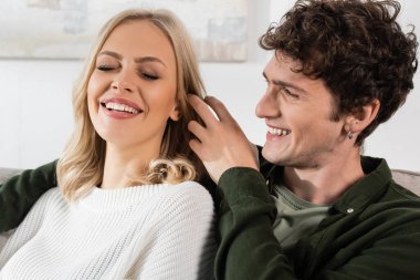 happy and curly man in shirt adjusting blonde hair of pretty girlfriend in white sweater 