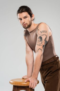 Long haired tattooed man standing near chair isolated on grey 