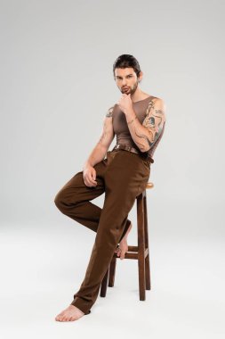 Full length of stylish tattooed man touching chin while sitting on grey background