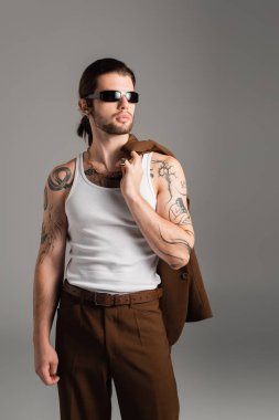 Tattooed man in sunglasses holding jacket isolated on grey 