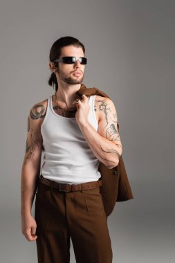 Tattooed man in sunglasses holding jacket isolated on grey  clipart