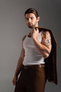 Long haired tattooed man holding brown blazer isolated on grey 