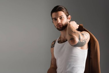 Portrait of stylish tattooed model in tank top holding jacket isolated on grey 