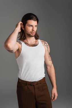 Stylish long haired man in tank top touching hair isolated on grey 