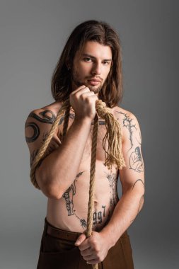 Muscular and tattooed model looking at camera while holding rope isolated on grey 
