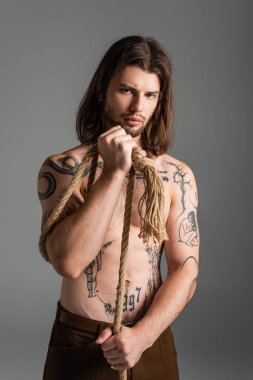 Shirtless tattooed man holding rope and looking at camera isolated on grey 