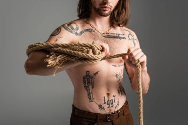 Cropped view of long haired model with tattoo on body holding rope isolated on grey 