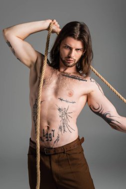 Shirtless tattooed model posing with rope and looking at camera isolated on grey 
