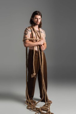 Full length of tattooed man with rope on body on grey background 