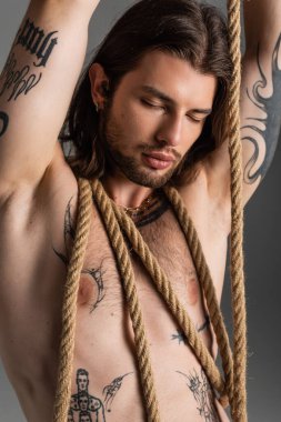 Long haired and tattooed model posing with rope isolated on grey 