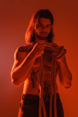 Sexy tattooed man holding rope isolated on red with light 