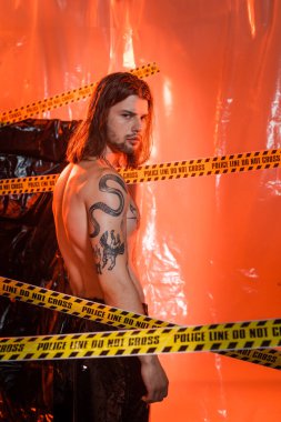 Shirtless tattooed model looking at camera near police line and cellophane at background 