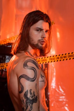 Portrait of shirtless tattooed man looking at camera near blurred police line and polyethylene