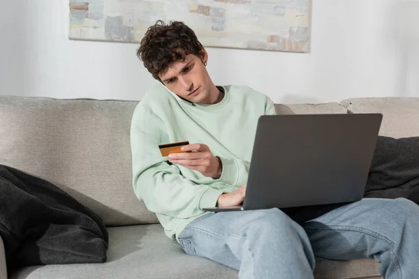 stock image curly young man holding credit card while talking on smartphone near laptop 