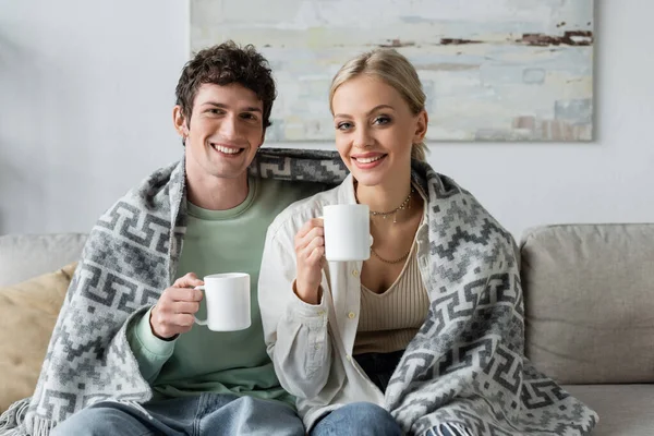 happy young man and smiling blonde woman covered in blanket holding cups of tea while sitting on couch 