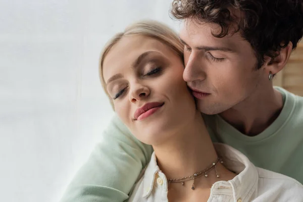 stock image curly man in sweatshirt hugging and kissing cheek of young girlfriend with closed eyes 