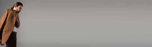 Tattooed and bearded man holding brown blazer isolated on grey, banner 